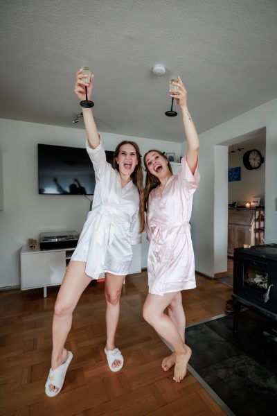 Zwei Frauen in Morgenmänteln jubeln mit Sektgläsern beim Getting Ready