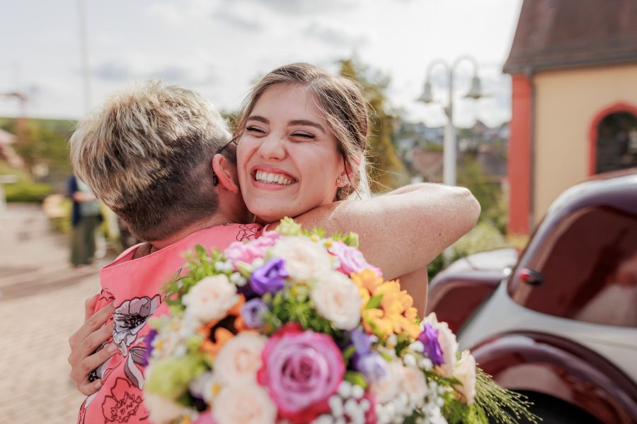 Die Braut umarmt lachend einen Gast vor der Kirche